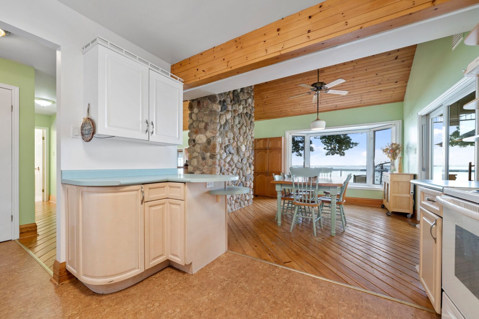 A kitchen with light green countertops, beige cabinets, and white appliances. In the next room, there is a dining table and chairs on hardwood floors.