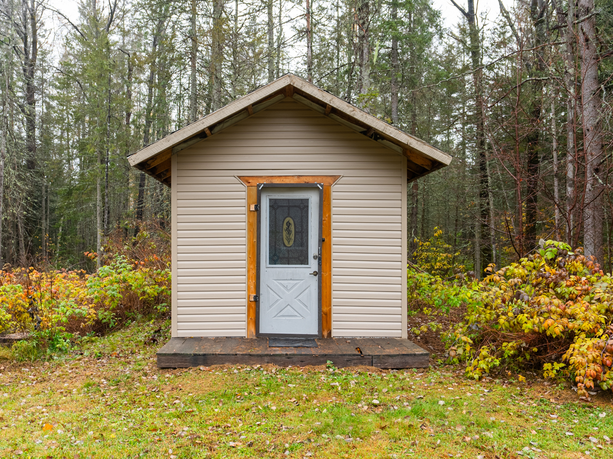 A small bunkie sits in front of the tree line on a grassy property.