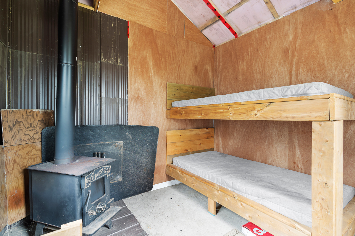 Interior of a small bunkie, with single bunk beds on one side and a wood stove on the other side.