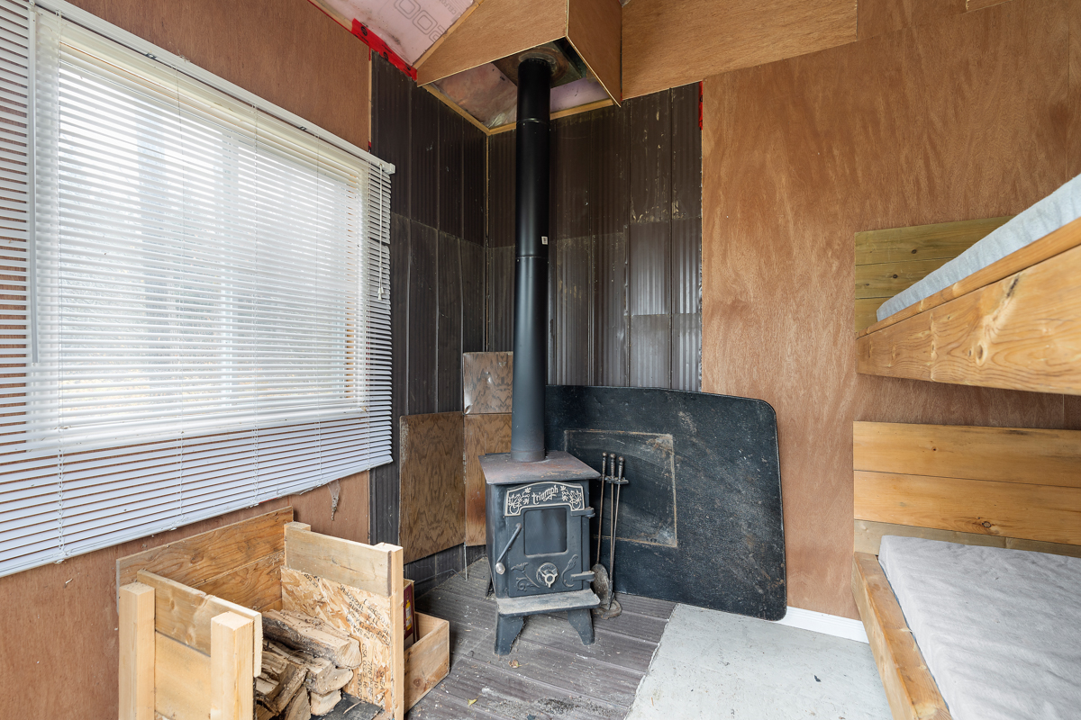 Interior of a small bunkie, with single bunk beds on one side and a wood stove on the other side. A big window lets in lots of natural light.