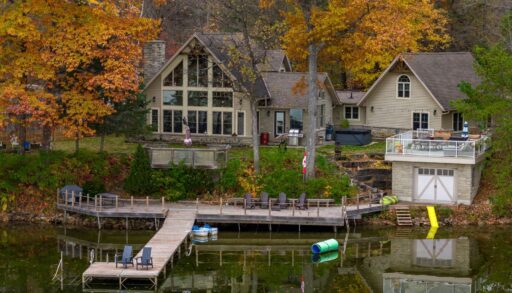 A large cottage with huge windows, a boathouse, and a long dock sits on the shore of a calm lake, surrounded by trees with colourful fall leaves.