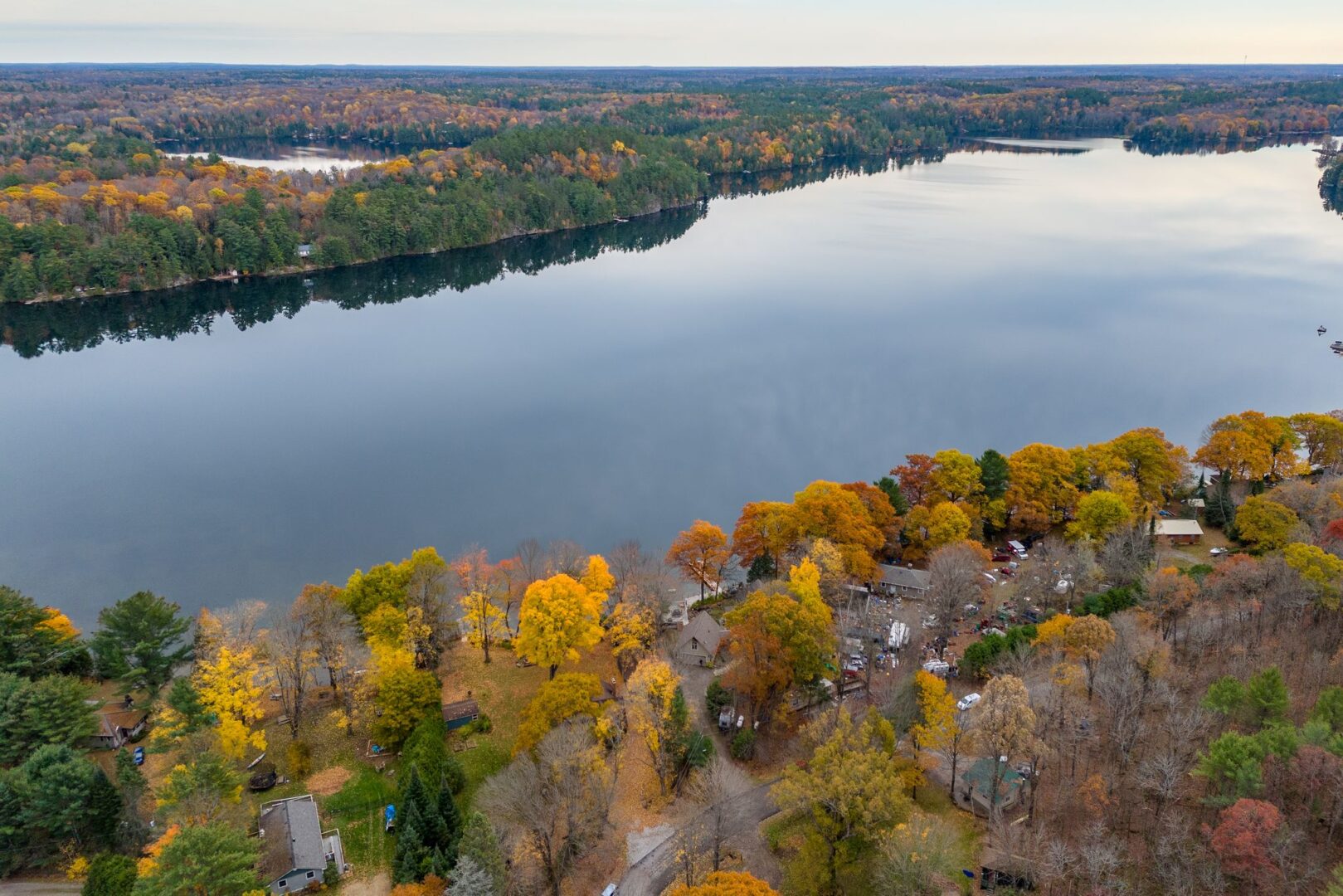 Overhead view of a calm lake. Trees with colourful fall leaves line the shore on either side, and some big cottages sit amongst the trees near the water..