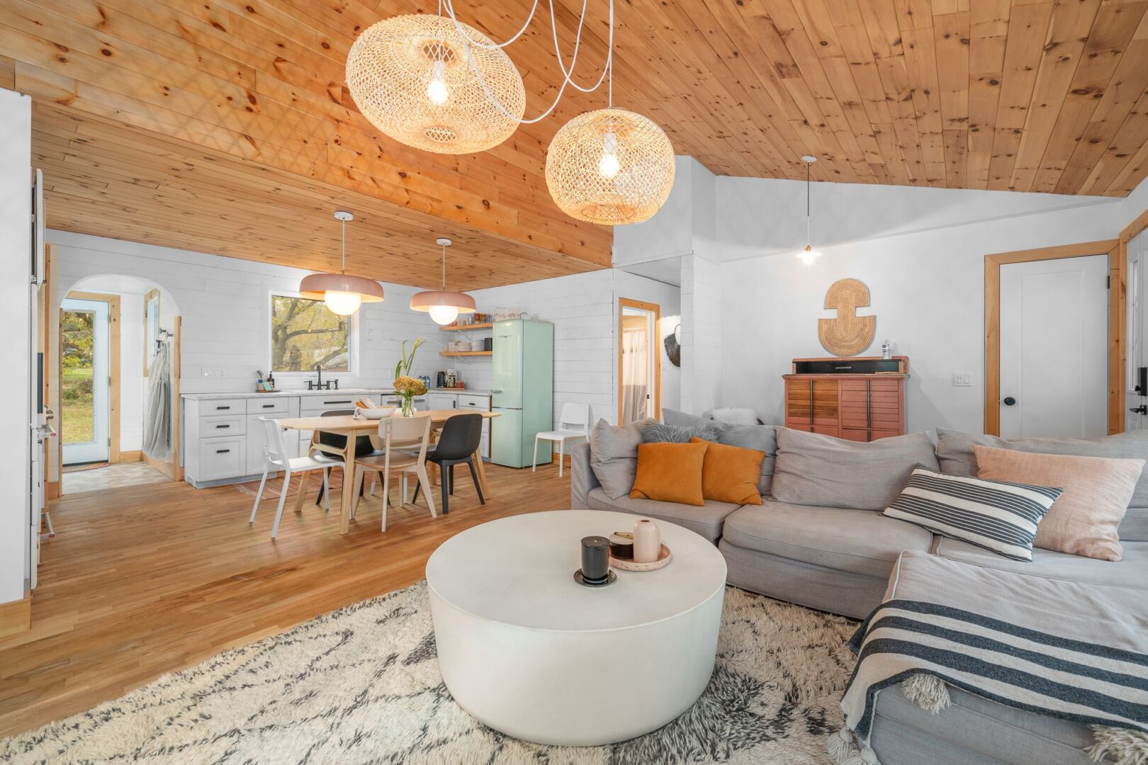 A bright, modern interior of an open-concept living area with wood-panelled ceilings, white and light grey furniture, and a dining table..