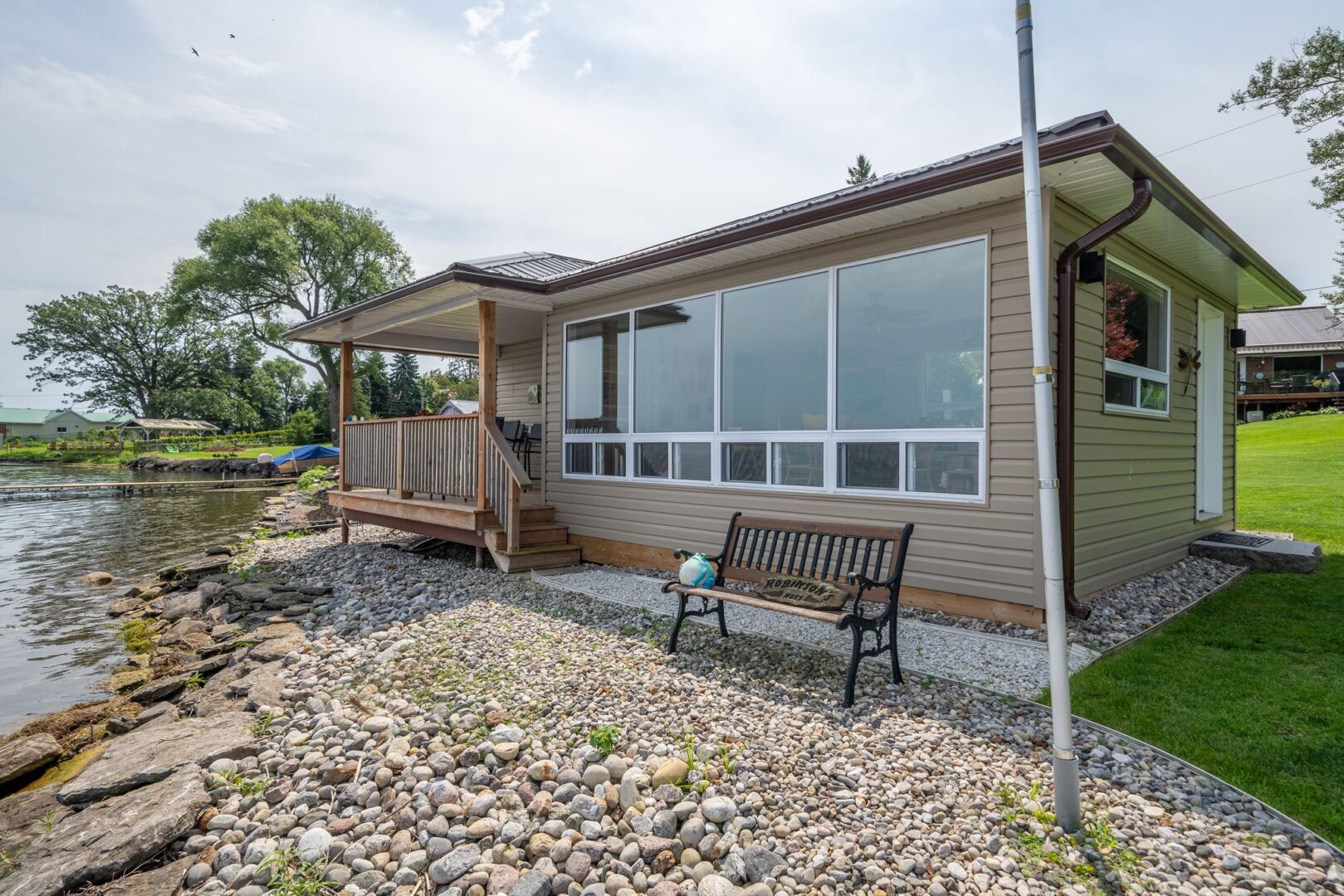 The back of a big bunkie on a rocky shoreline. A big window spans the lake-facing wall of the bunkie, and stairs lead up to a deck on the other side..