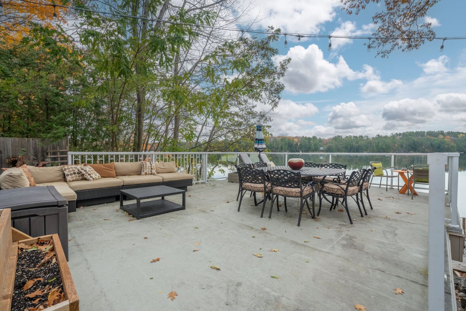 A large patio spans the roof of a boathouse, looking out over a lake. There is outdoor furniture, including a dining table and a couch, on the patio.