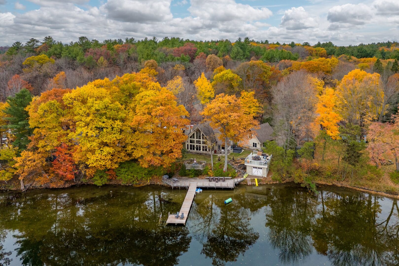 A large cottage with huge windows, a boathouse, and a long dock sits on the shore of a calm lake, surrounded by trees with colourful fall leaves.