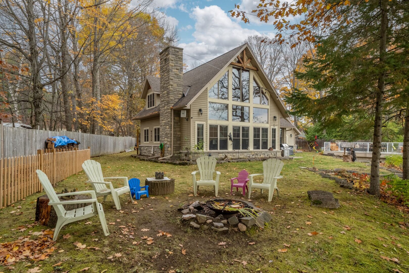 Colourful Muskoka chairs surround a fire pit in front of a big, luxurious cottage.