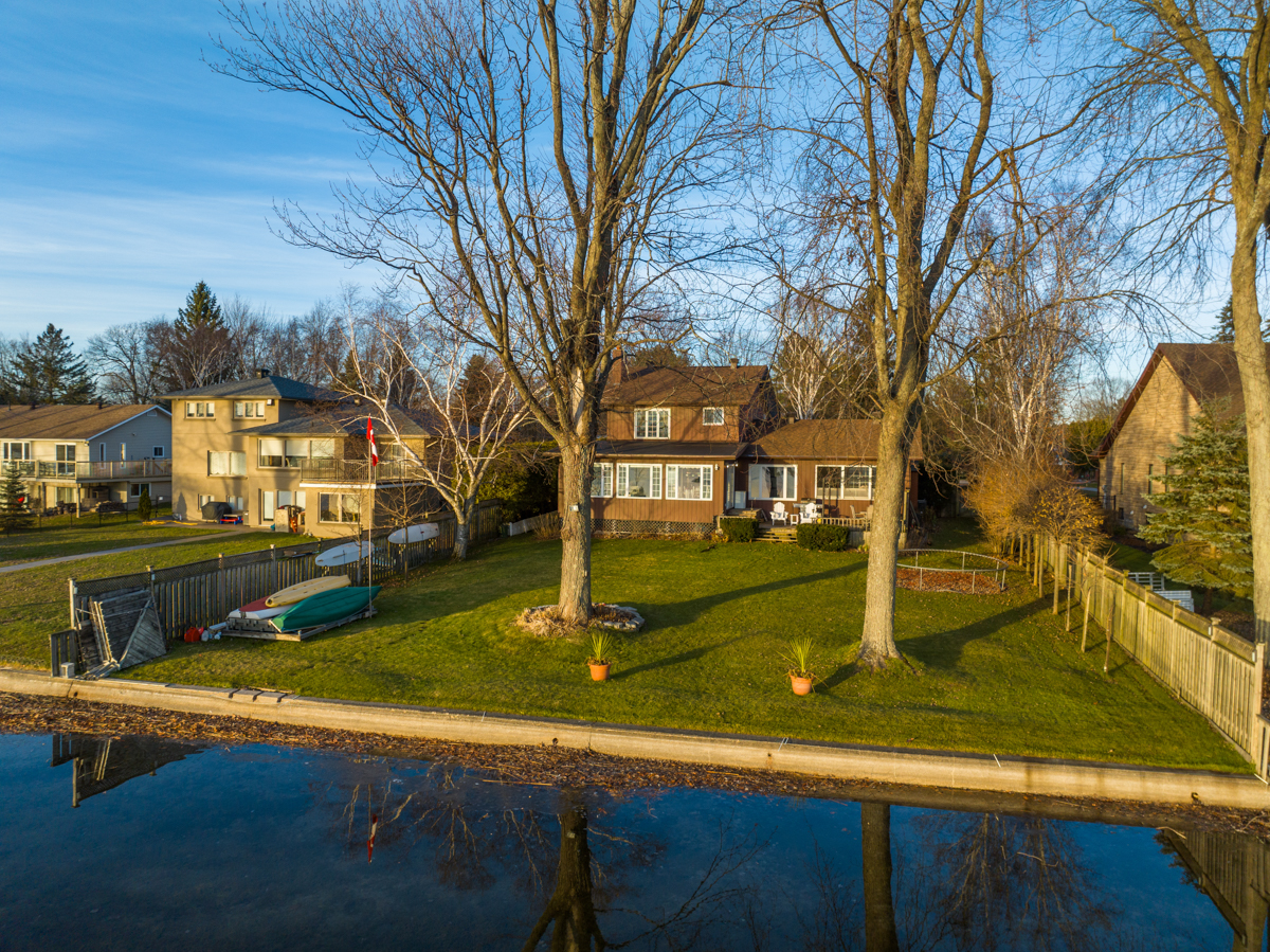 A grassy lot backs onto the shore of a calm lake in a residential area. A two-story home and two trees sit on the lot.
