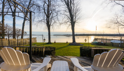 View from two white Muskoka chairs on a raised deck, looking out across a big grassy area and a lake beyond.