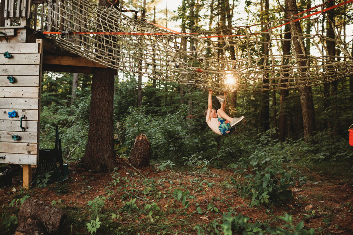 A young boy hanging from the net of a jungle gym, thick woods and the setting sun in the background