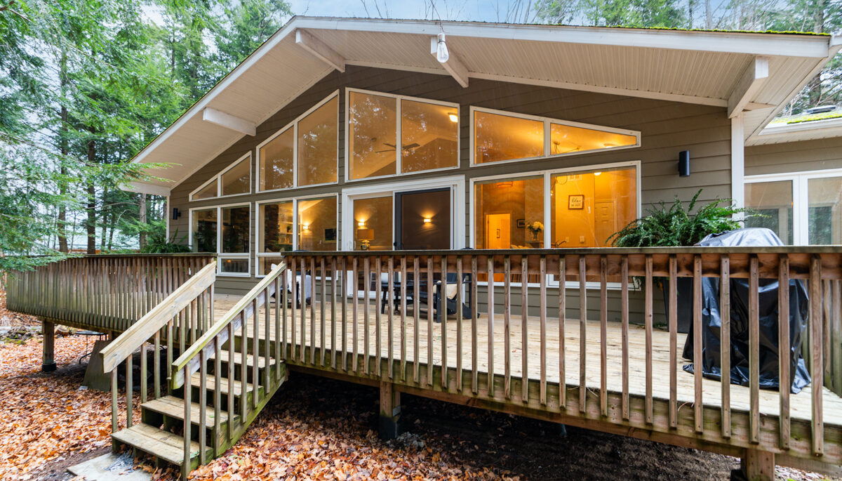 A cottage sits in a forested area. It has floor-to-ceiling windows, a gently peaked roof, and a large deck.