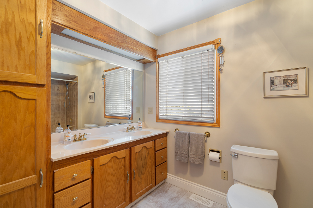 A three-piece bathroom with a toilet, double sinks, wooden cupboards, and a shower-tub combo.