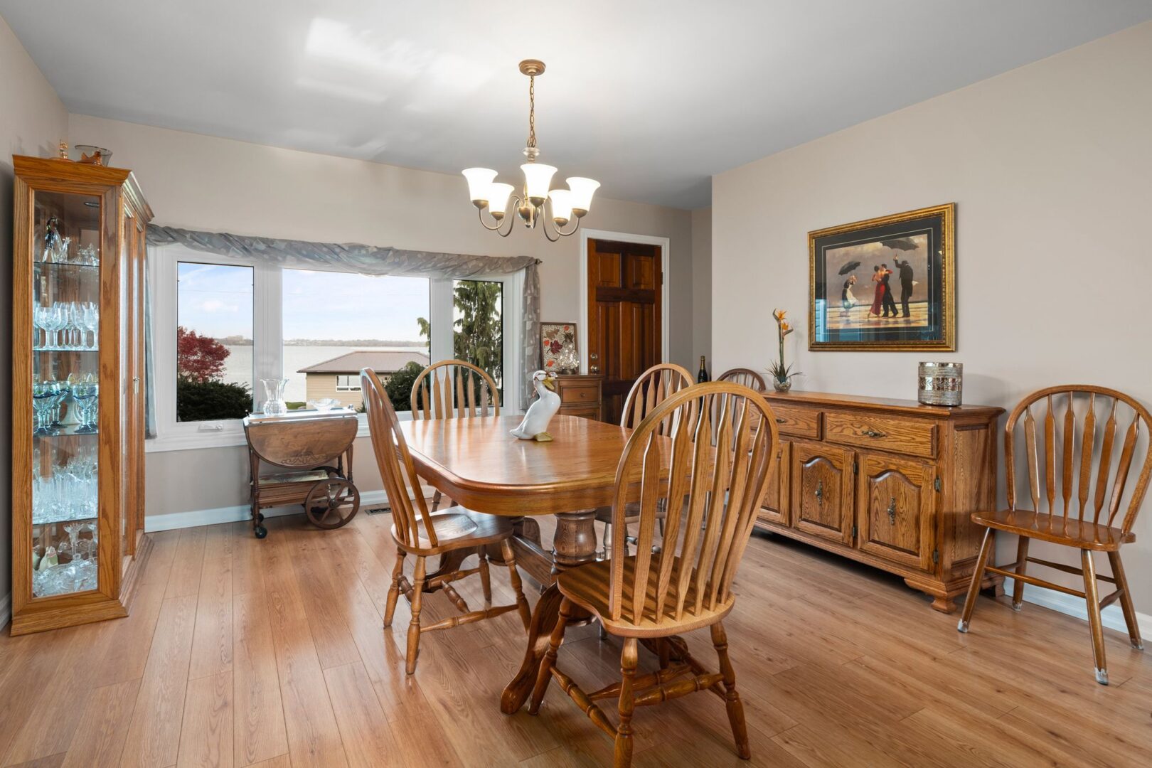 A bright room with hardwood flooring, a wooden table and chairs, and a big window that faces the water.