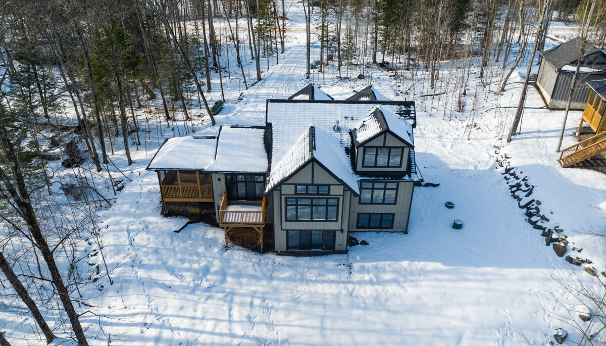 A large home sits in a clearing of a wooded, rural area. There is snow on the ground and the trees are bare.