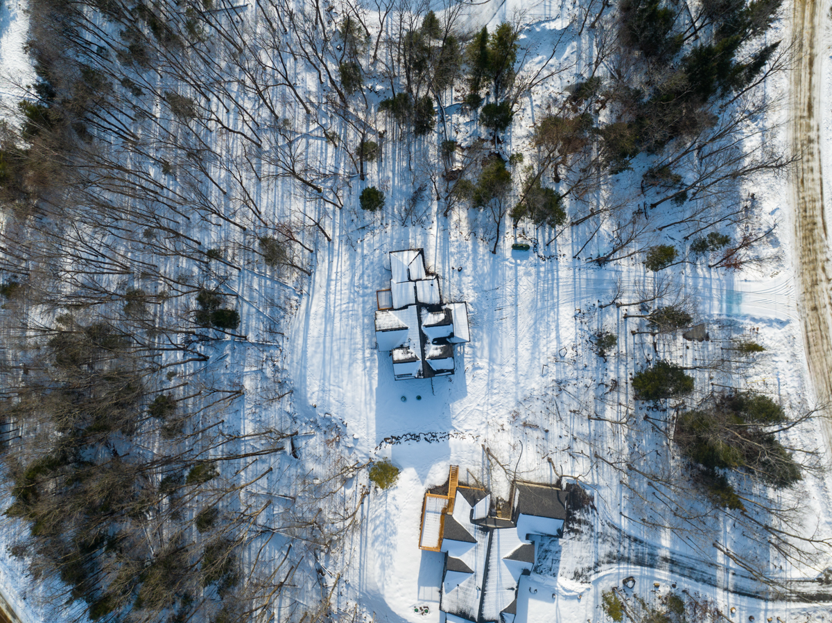 Aerial view of a large house on a rural property. There is snow on the ground, and bare trees surround the area of the house. One neighbouring house also sits among the trees.