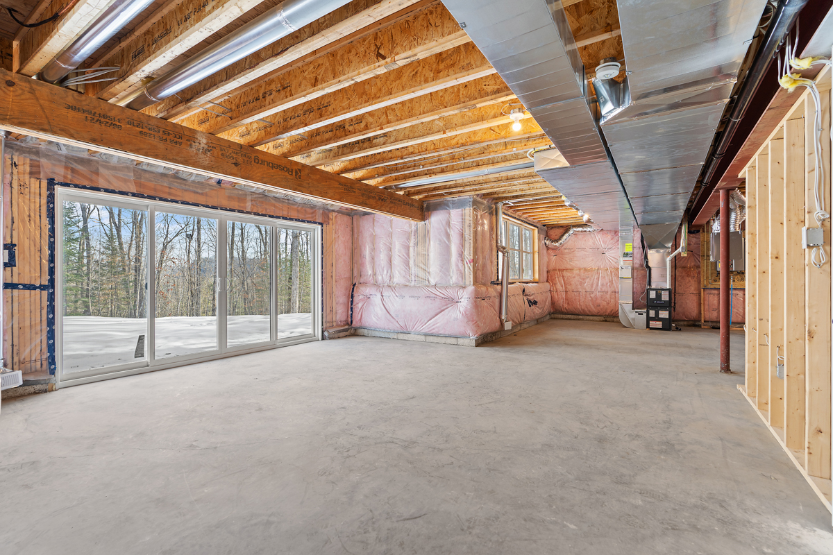 A large unfinished basement area with exposed wooden beams and insulation. Large glass doors walk out to an outdoor space, letting in lots of natural light.