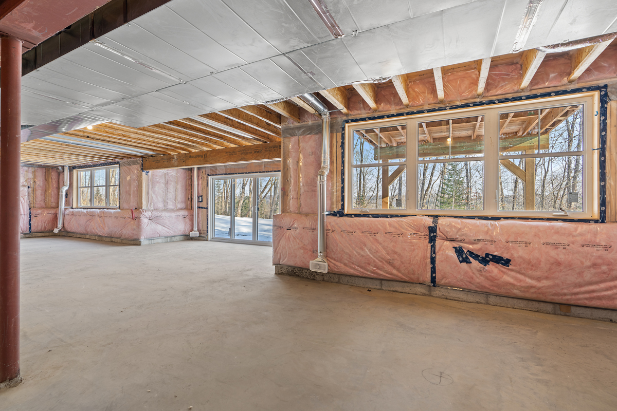 A large unfinished basement area with exposed wooden beams and insulation. Large glass doors walk out to an outdoor space, letting in lots of natural light.
