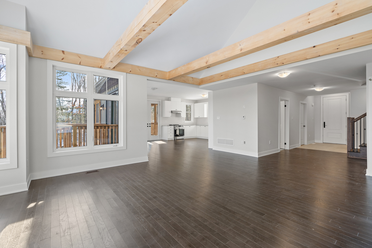 A bright, empty space with dark hardwood floors, white walls, and exposed beams of light wood spanning a white vaulted ceiling. The area has a large window and flows into a kitchen space.
