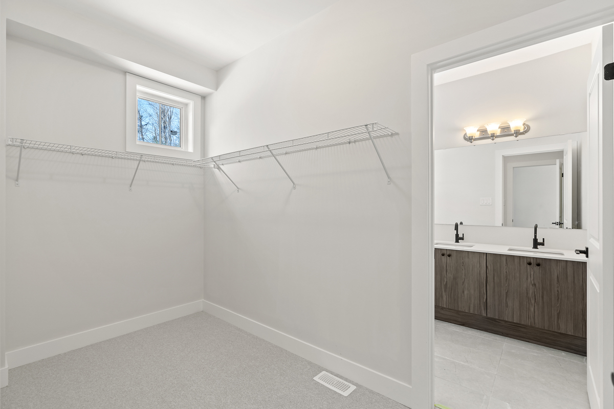 A big, empyt walk-in closet with two build-in hanging racks, a small window, white walls, and light gray carpet flooring. A doorway leads to a bathroom.