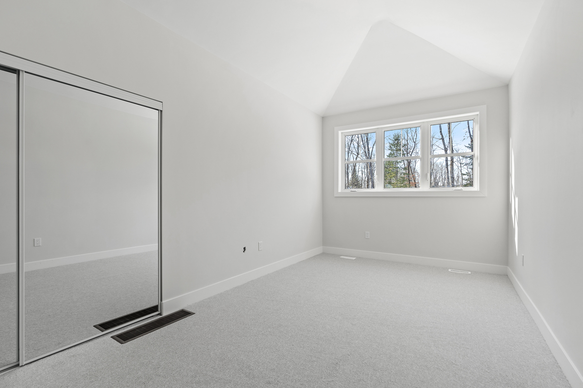 An empty bedroom with a big window, white walls, a white vaulted ceiling, and light gray carpet flooring. There is a big closet with sliding mirror doors on one wall.