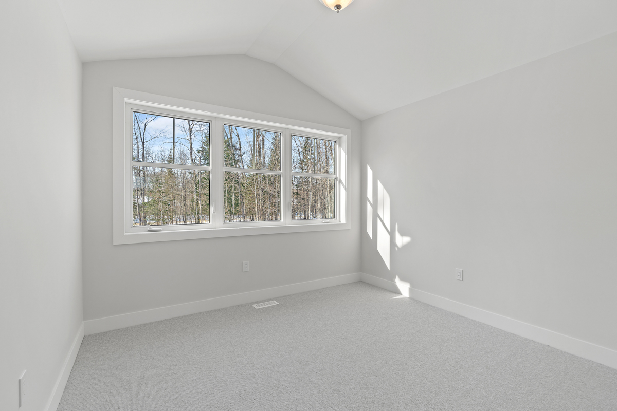 An empty bedroom with a big window, white walls, a white vaulted ceiling, and light gray carpet flooring.