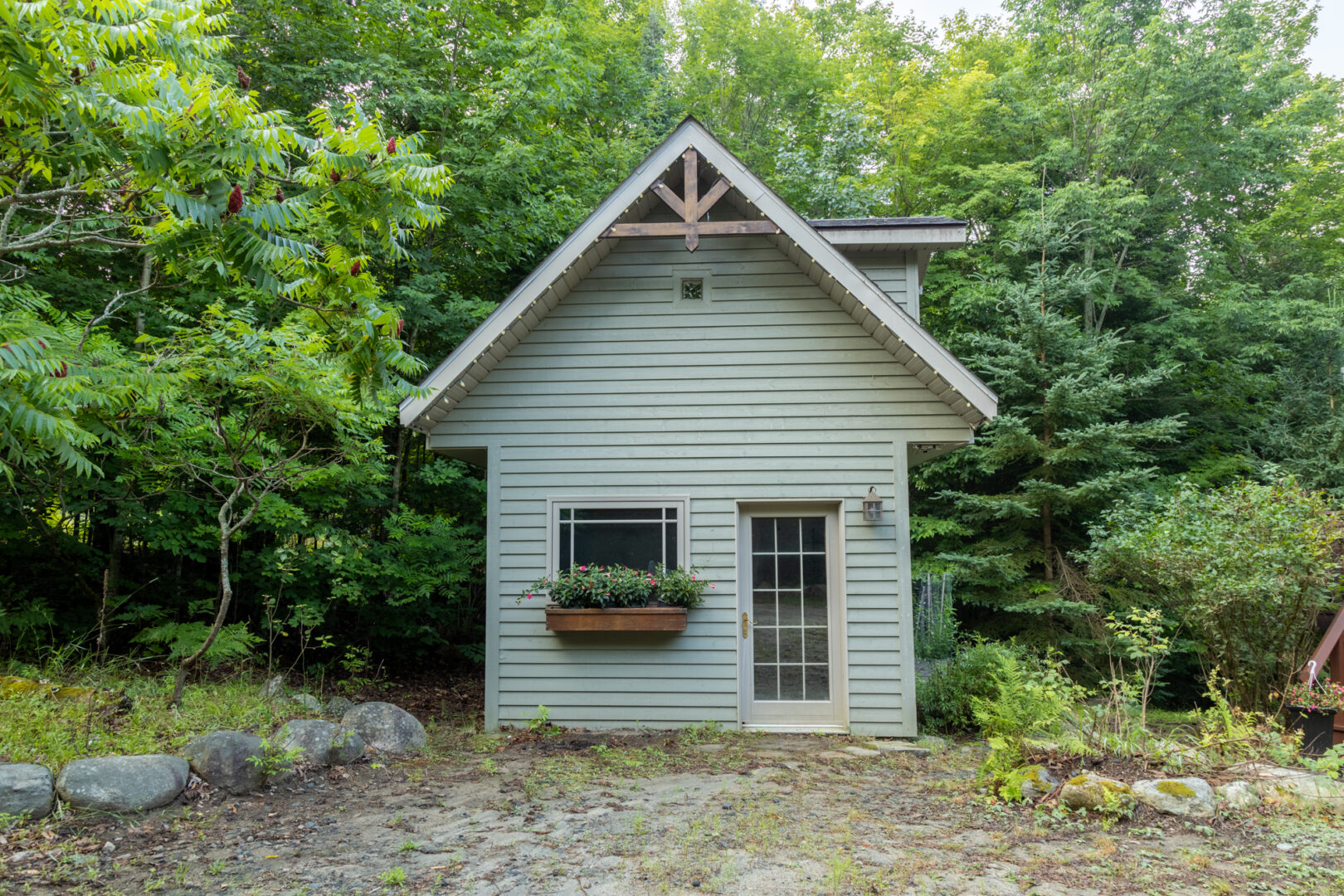A small shed with a green-gray exterior, a peaked roof, a window, and a door.