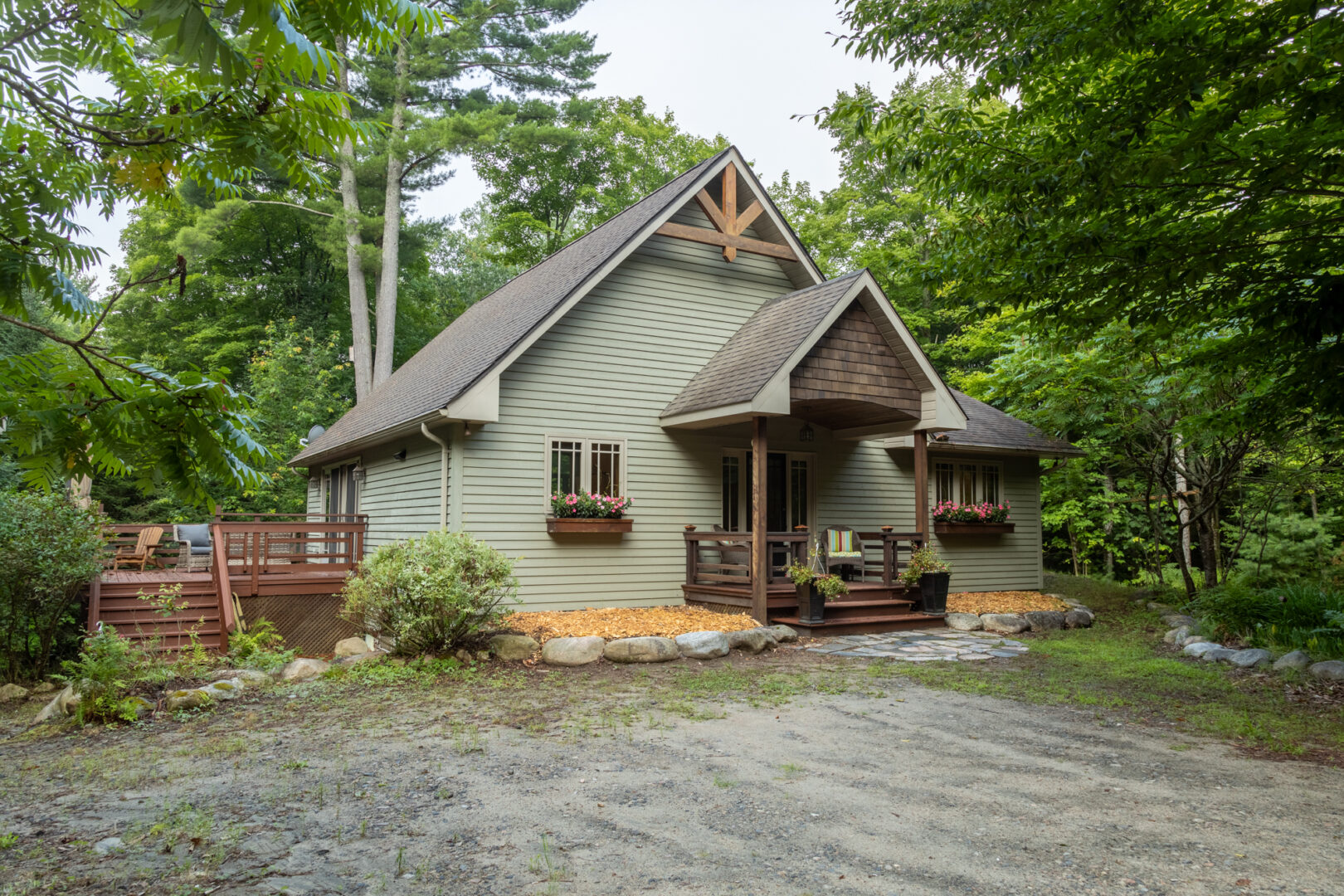 A cottage with light green siding, a slanting roof, and a big side deck sits on a dirt and gravel driveway, surrounded by green trees.