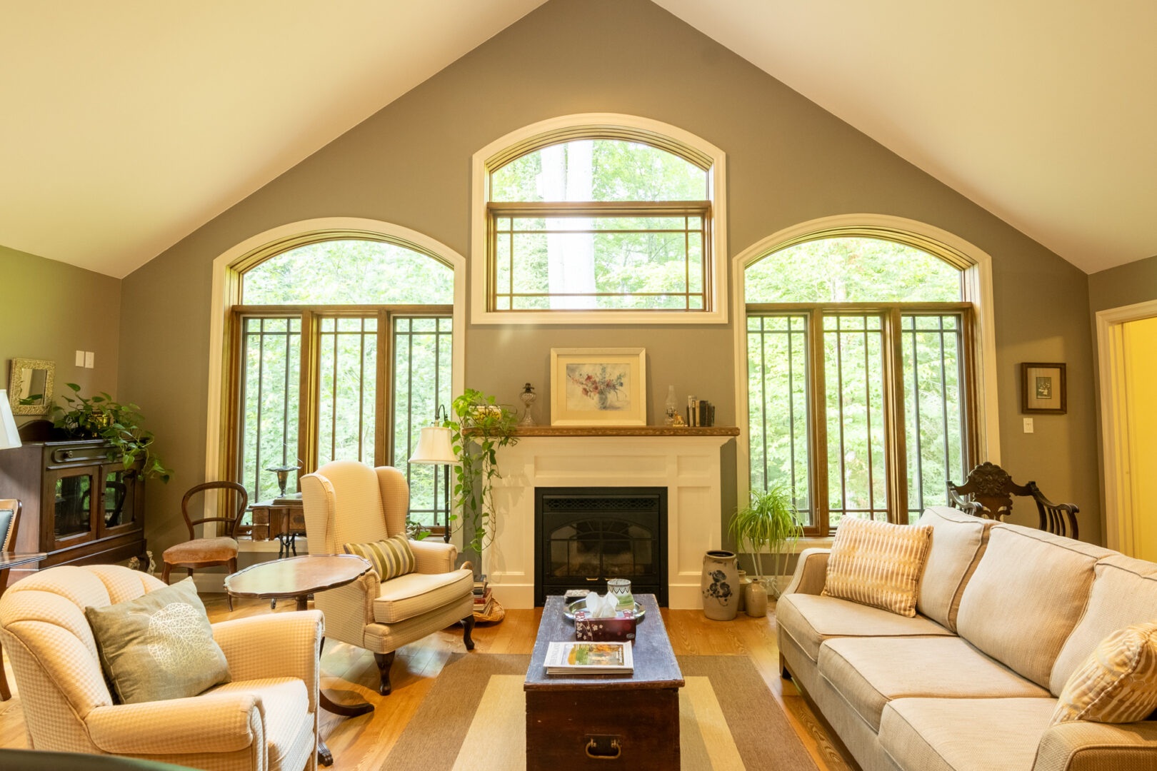 A bright but cozy living space with green-gray painted walls, high, slanted ceilings, and large windows. A fireplace sits in the middle of the exterior wall, and there is a couch and two chairs in the middle of the room.