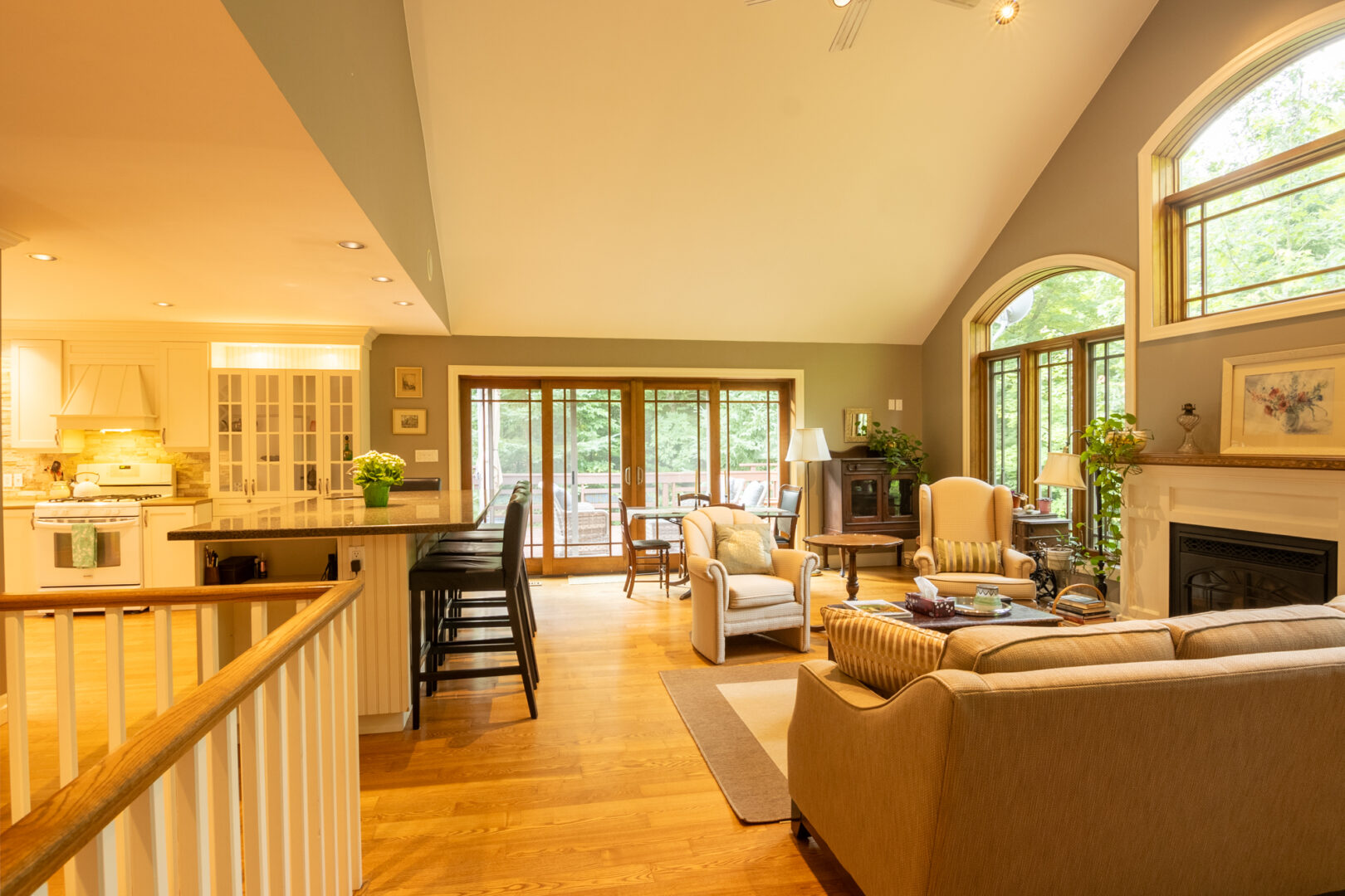 An open-concept main floor space that flows from a kitchen into a living area. The space has hardwood floors, a high, slanting ceiling, and double glass doors that open out to an outdoor deck area.