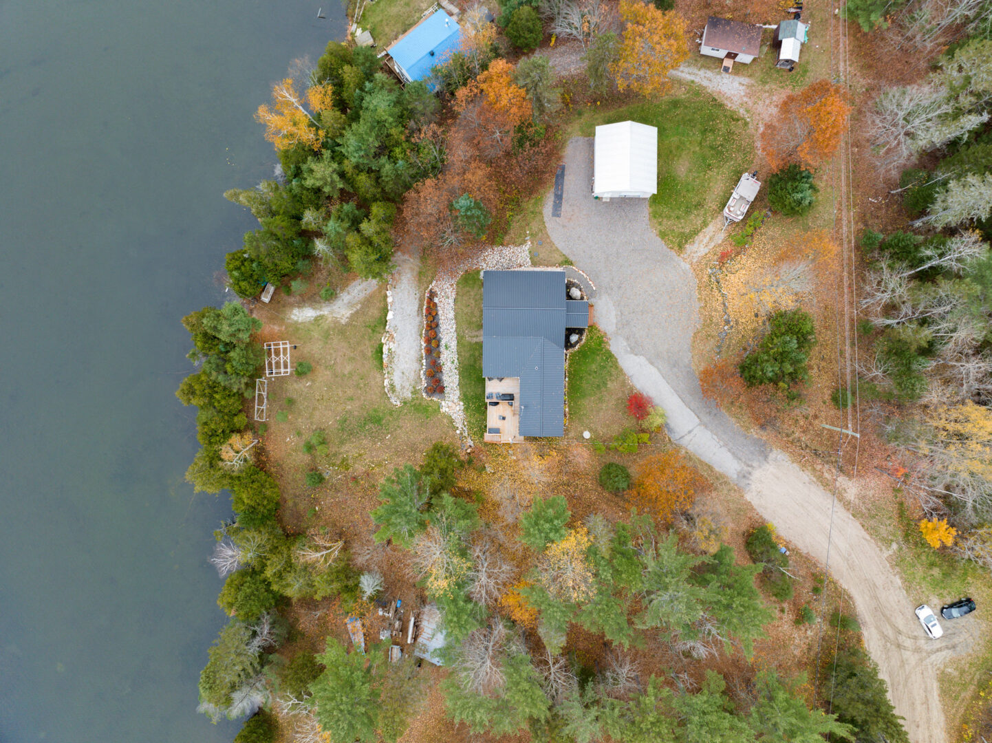 Aerial view of a small bungalow with a blue-gray roof, sitting waterfront on a calm lake. The front of the house has a large paved driveway, and the home is surrounded by trees and greenery.