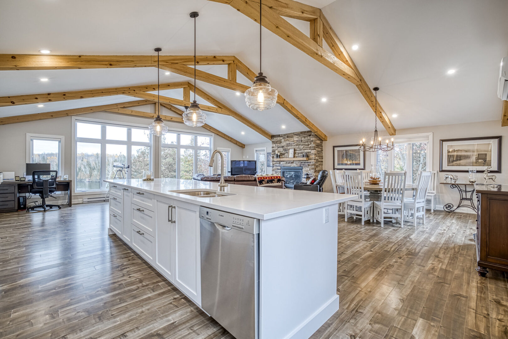A large, bright kitchen area with hardwood floors and high, cathedral-style ceilings with wooden beams. A large island sits in the middle of the bright space.