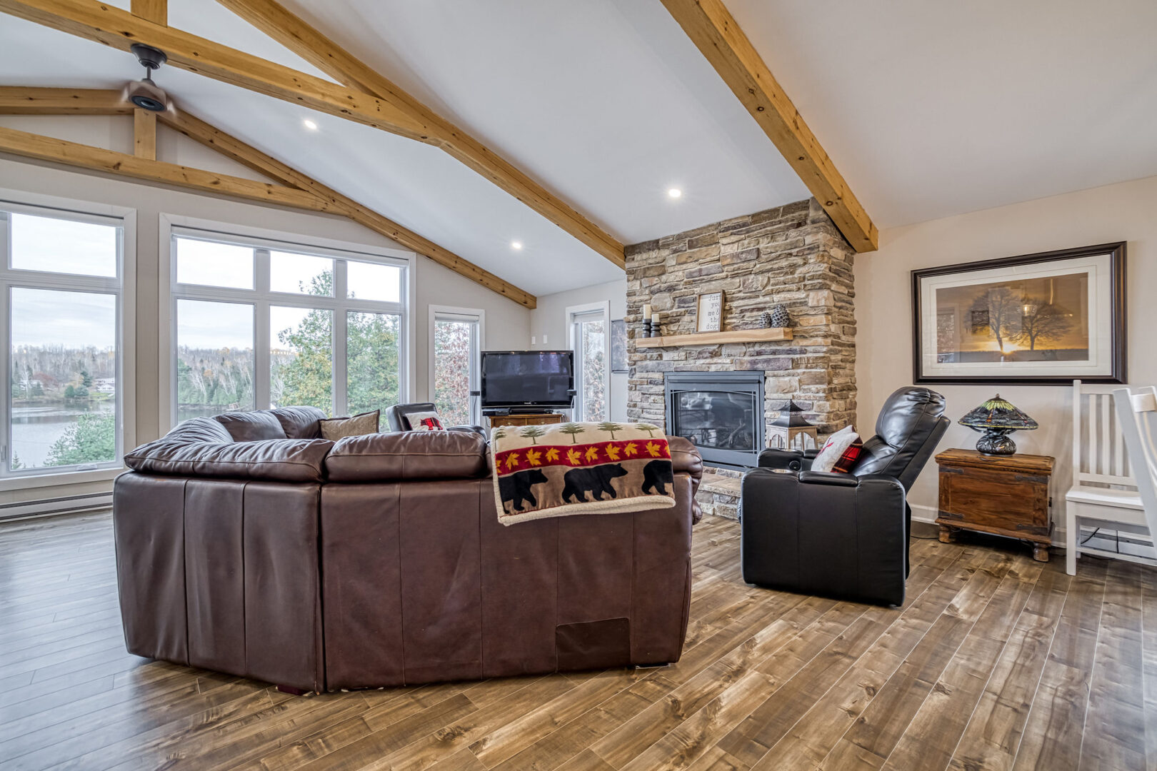 A bright living space with high ceilings, hardwood floors, a couch, a chair, big windows, and a floor-to-ceiling stone fireplace.