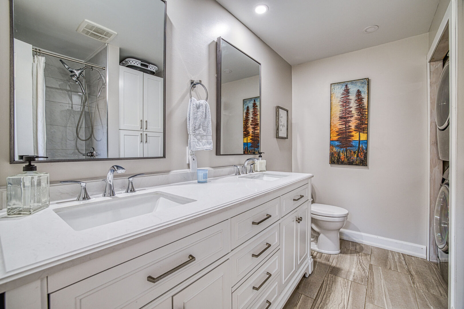A modern bathroom with white cabinetry, double sinks, hardwood flooring, a toilet, and a shower.