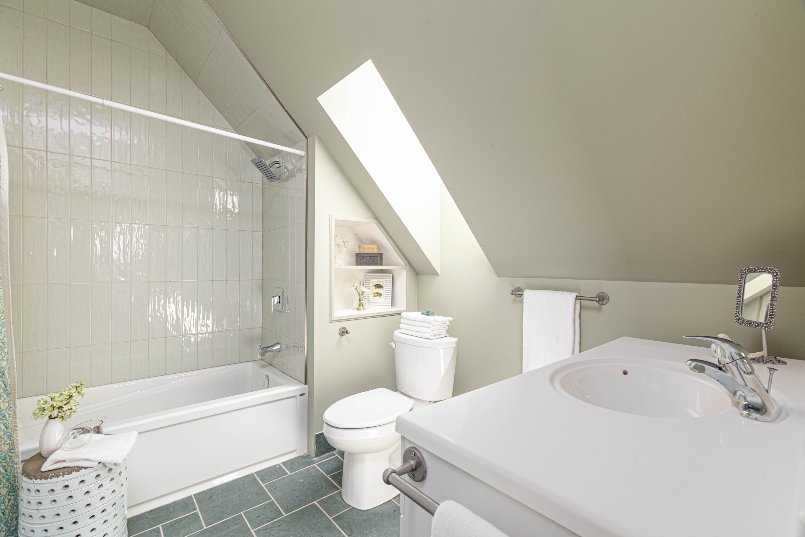 A big bathroom with a sloping ceiling, a skylight, a toilet, a sink, grey tile flooring, and a shower-tub combo.