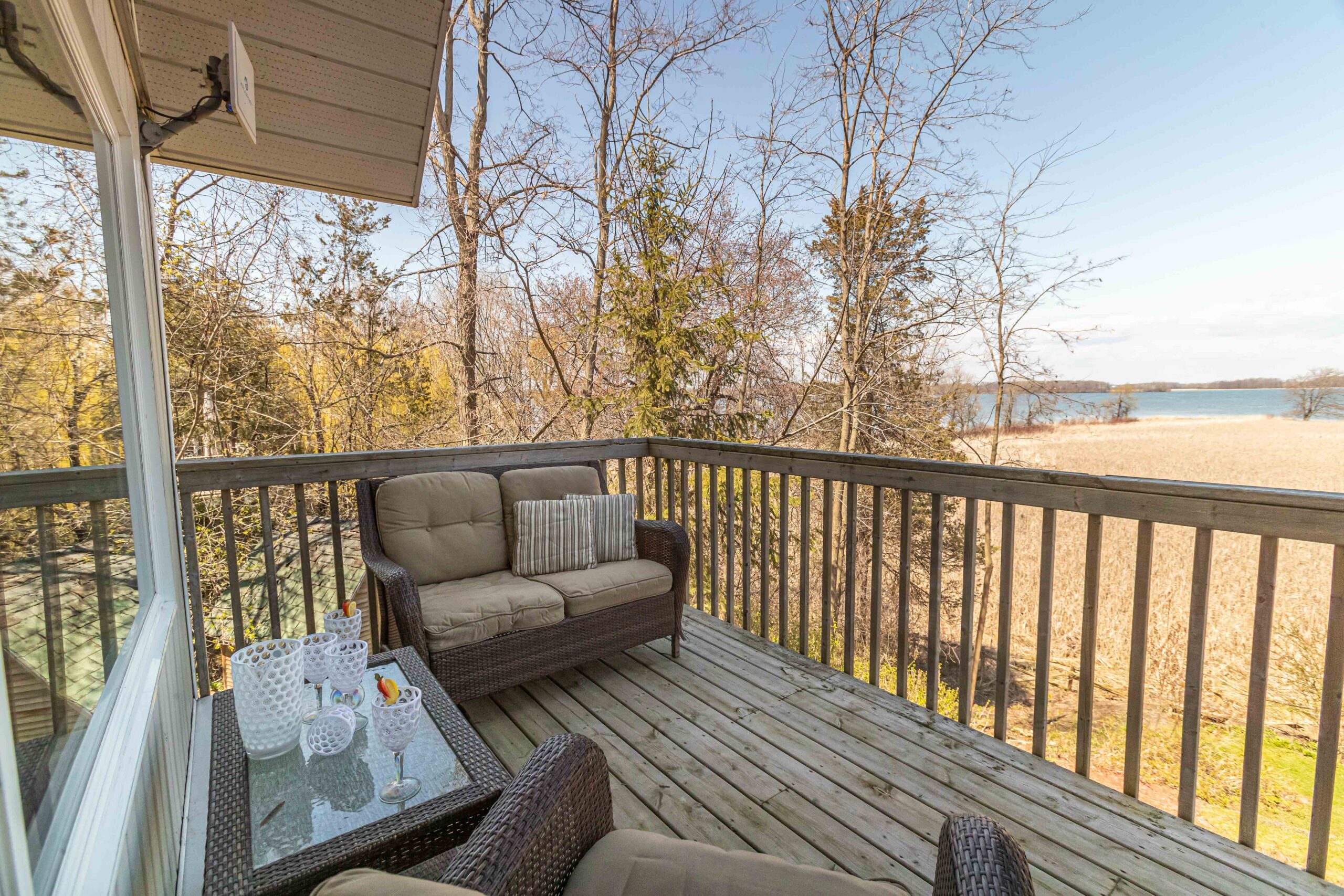 A wooden deck with outdoor seating, looking out over a lake with a marshy shoreline.