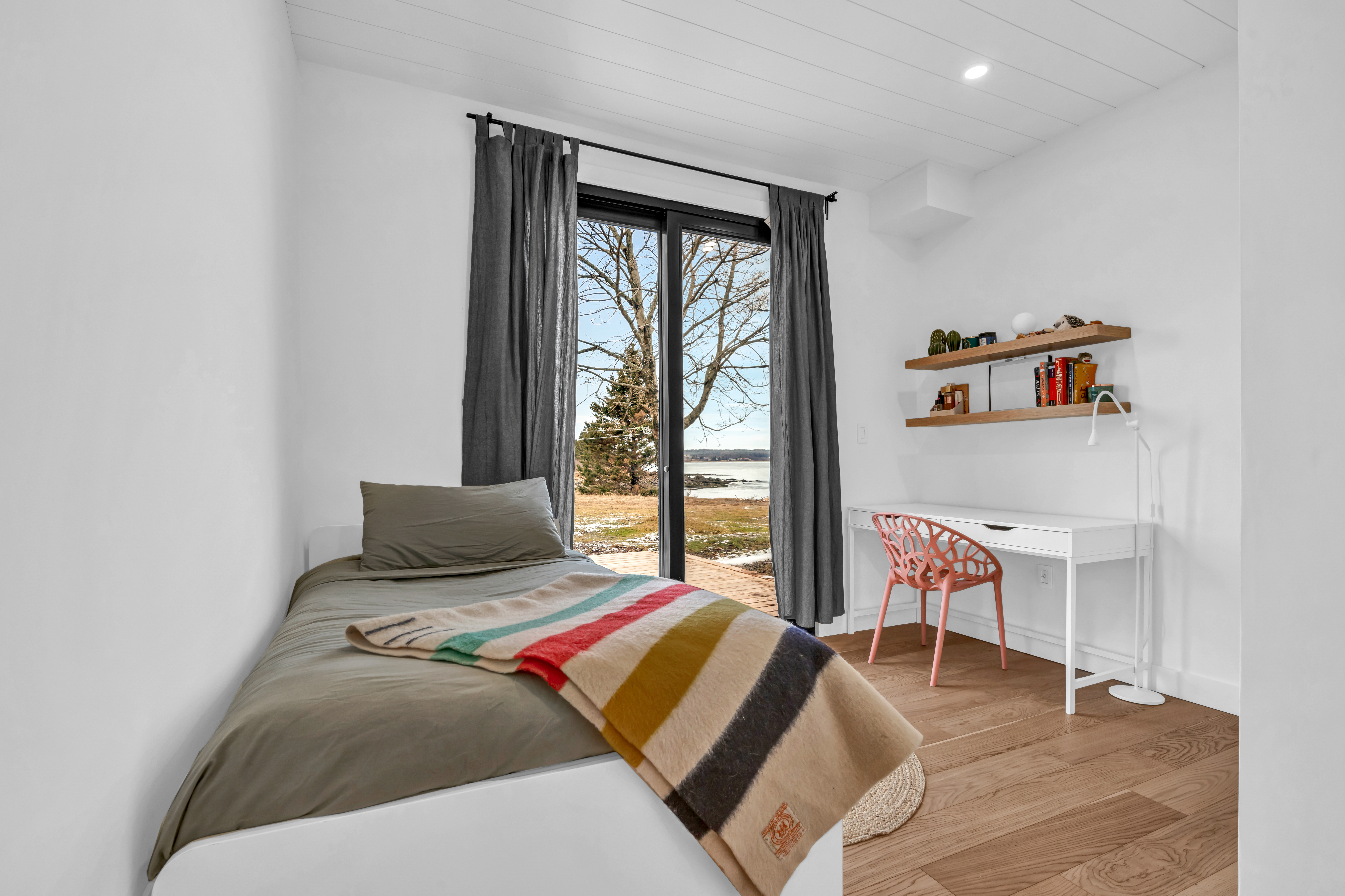 A simple bedroom with a small bed, a deck, hardwood flooring, and double glass doors that walk out to an outdoor deck.