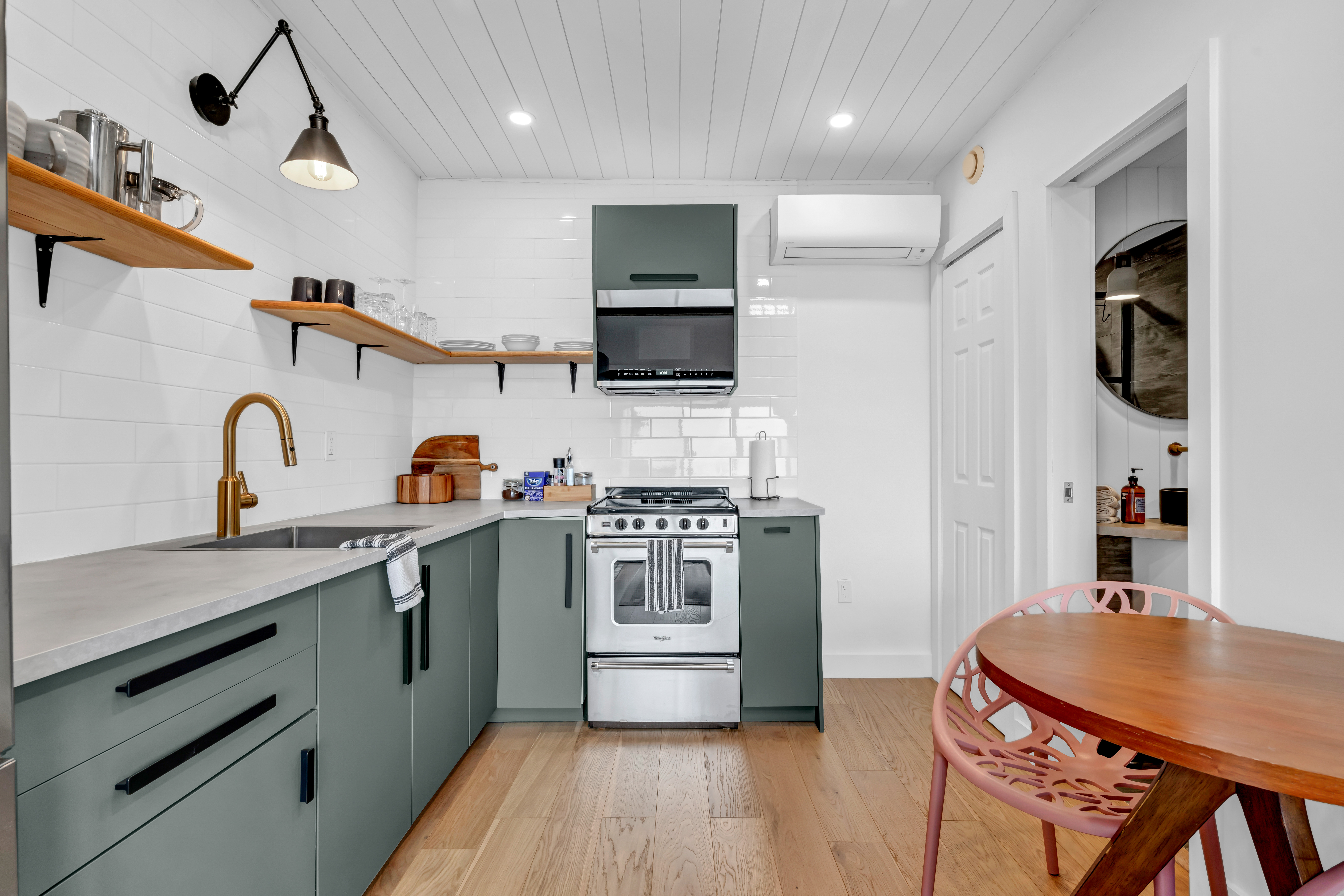 A modern kitchen with hardwood floors, green-gray cupboards, stainless steel appliances, and a small table and chairs.