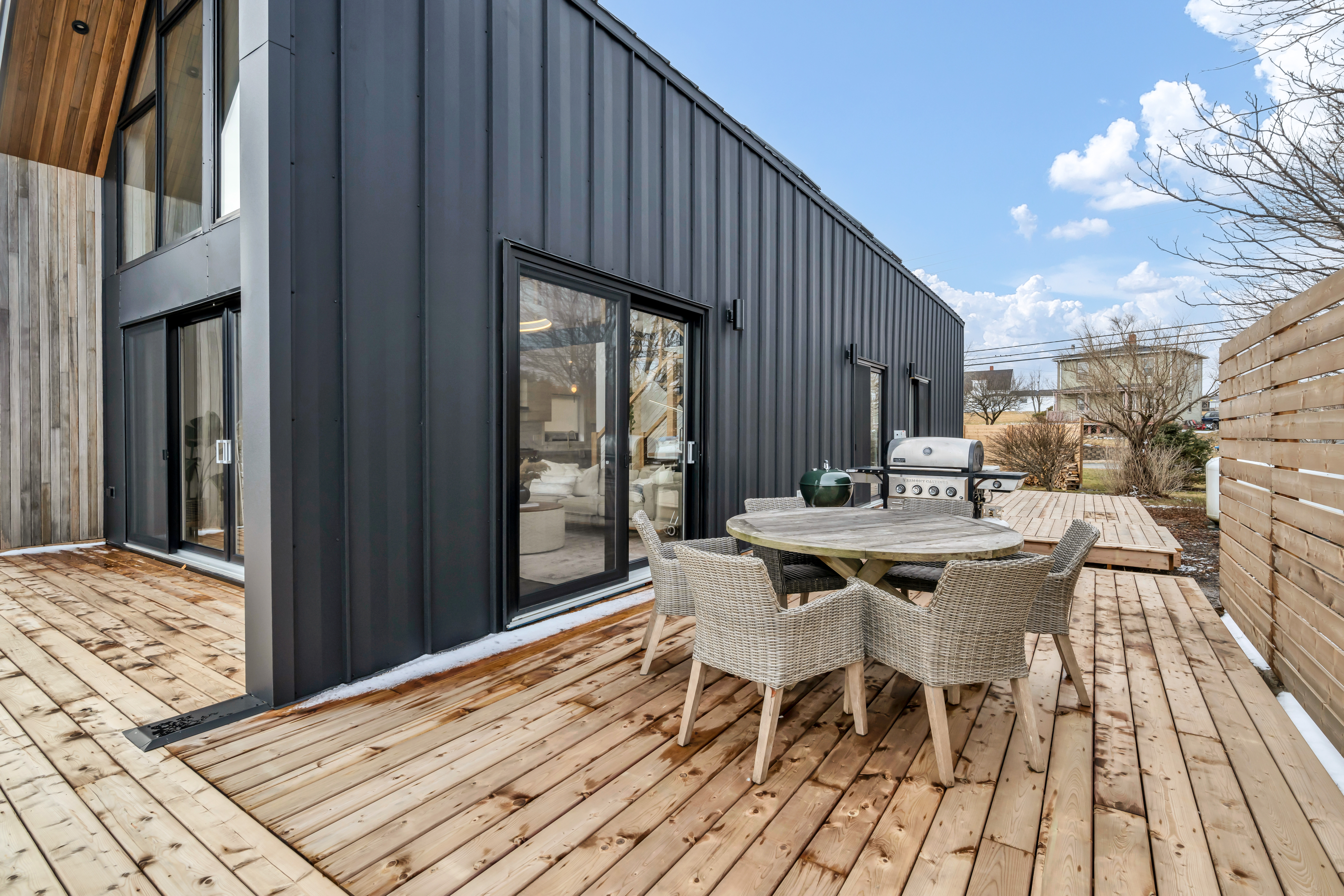 An outdoor dining table and chairs sit on a wooden deck off the side of an oceanfront home with big windows.
