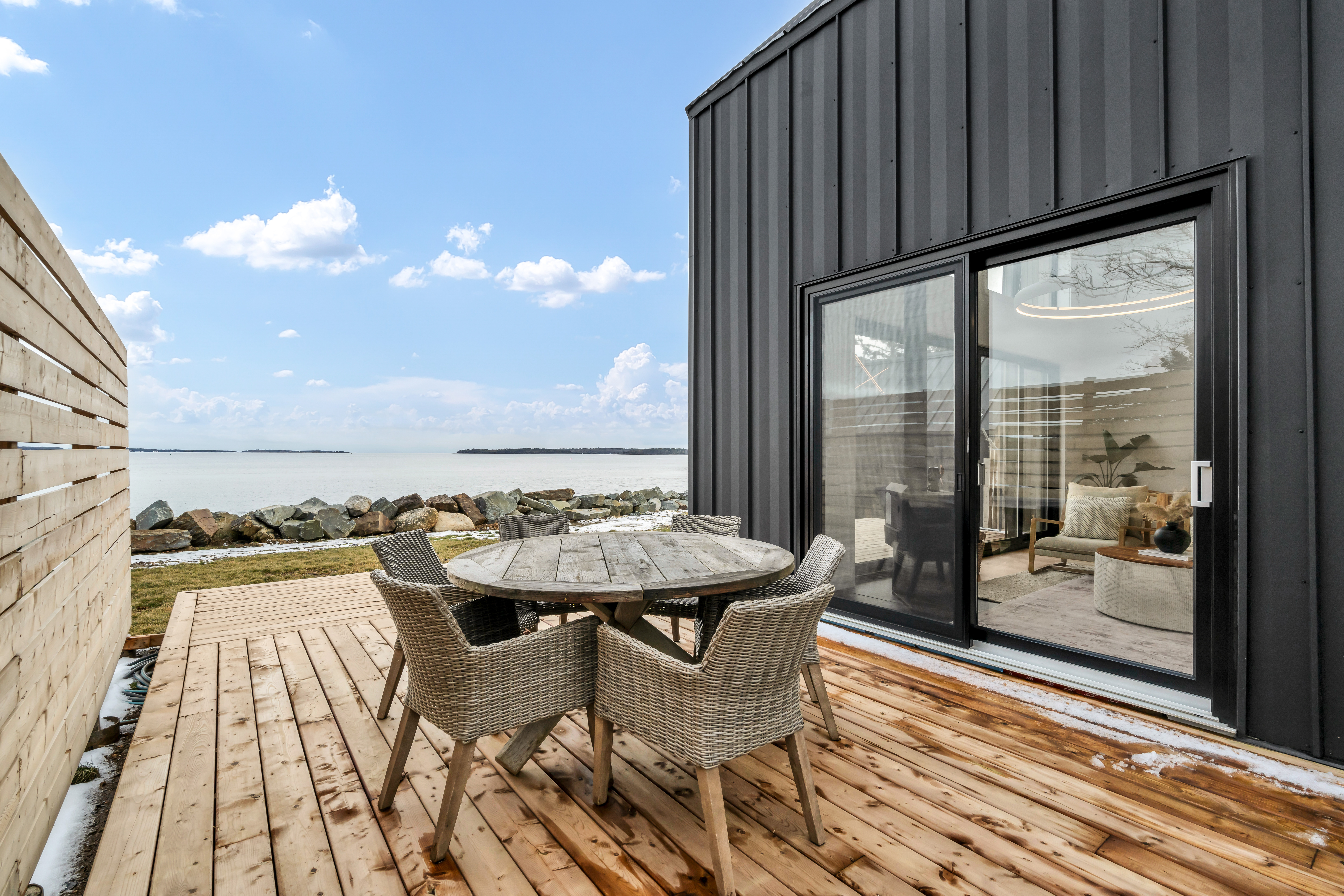 An outdoor dining table and chairs sit on a wooden deck off the side of an oceanfront home with big windows.