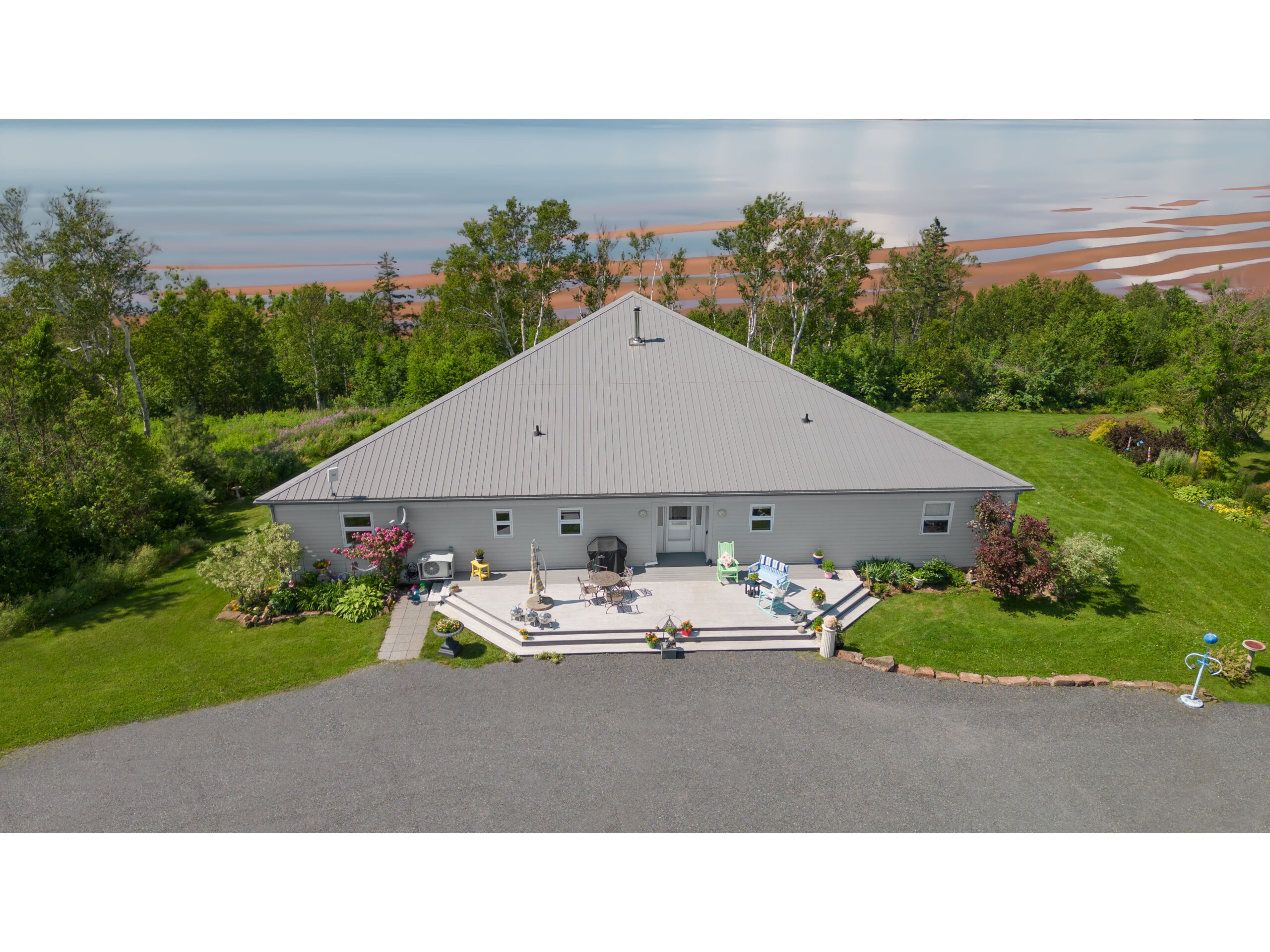 A large home with a big driveway, surrounded by trees and overlooking a red sand beach and a big stretch of water.