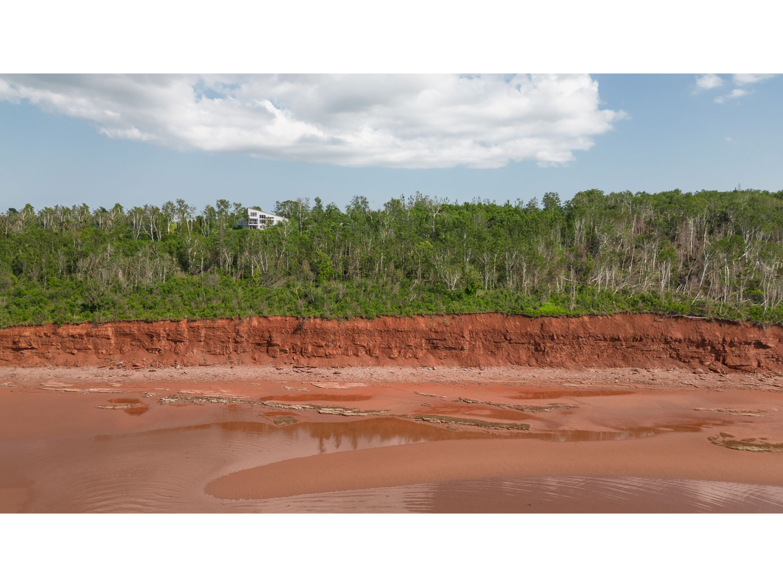 A red sand beach with green trees extending through the land behind. A big home is visible among the trees.