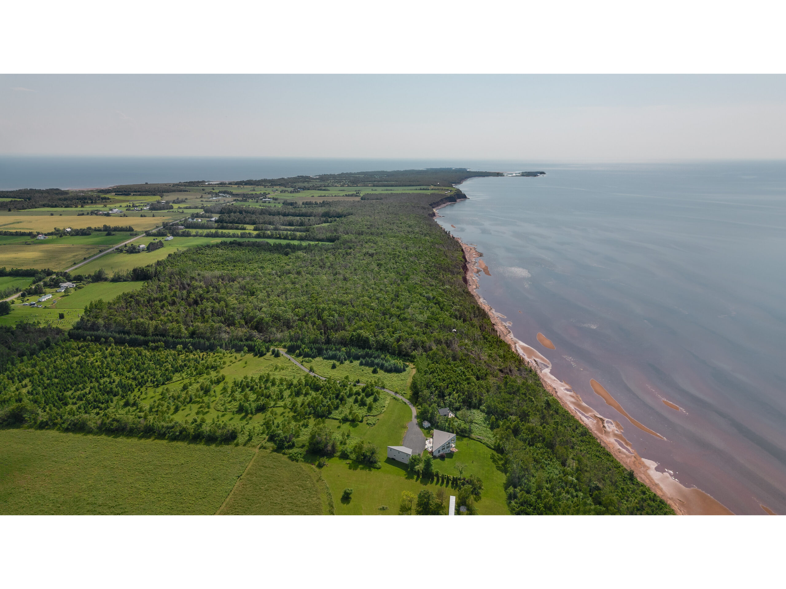 Overhead view of a stretch of waterfront land covered in green trees. The waterfront is red sand beach and calm water.