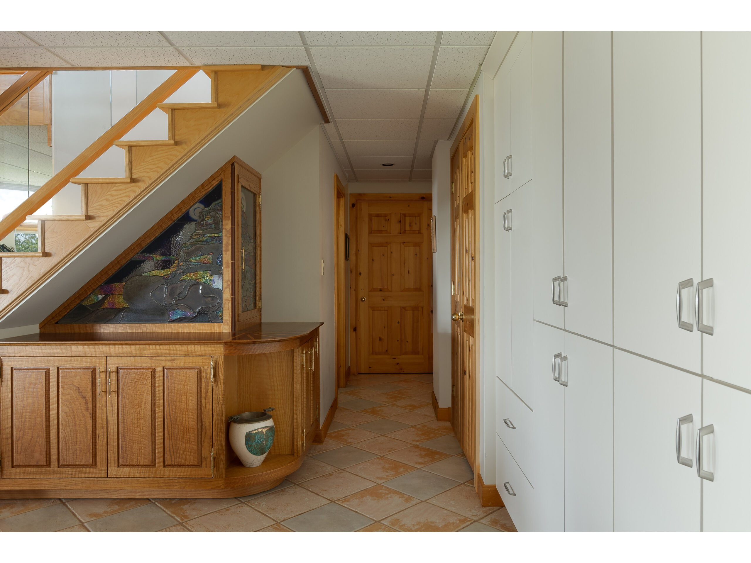 A downstairs hallway at the bottom of a staircase, with tiled flooring. Storage cupboards line the walls.