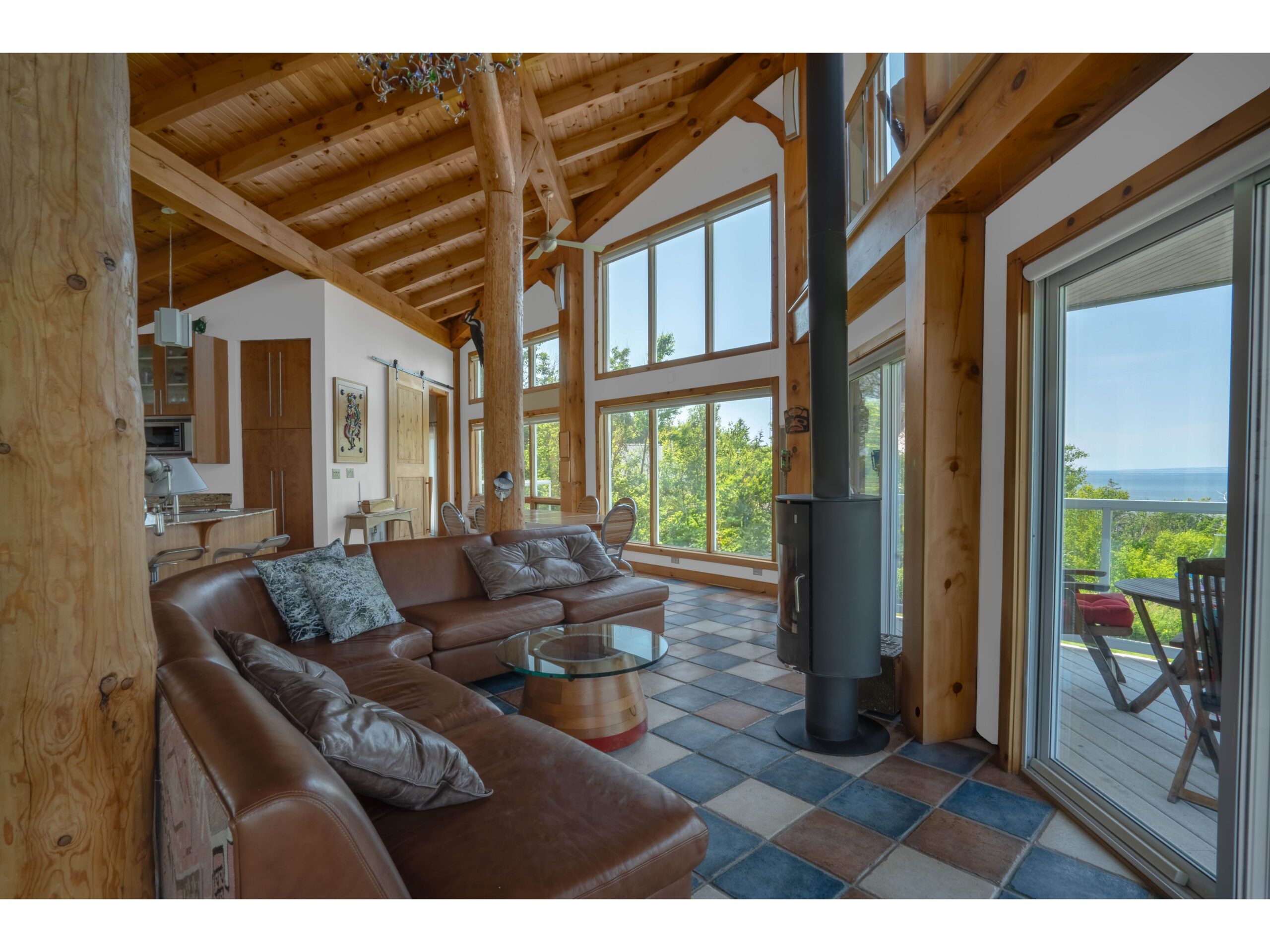 A large, bright, open-concept living area with colourful tiled flooring, wood accents, a leather couch and a wood stove. Big windows reveal green trees and water outside.