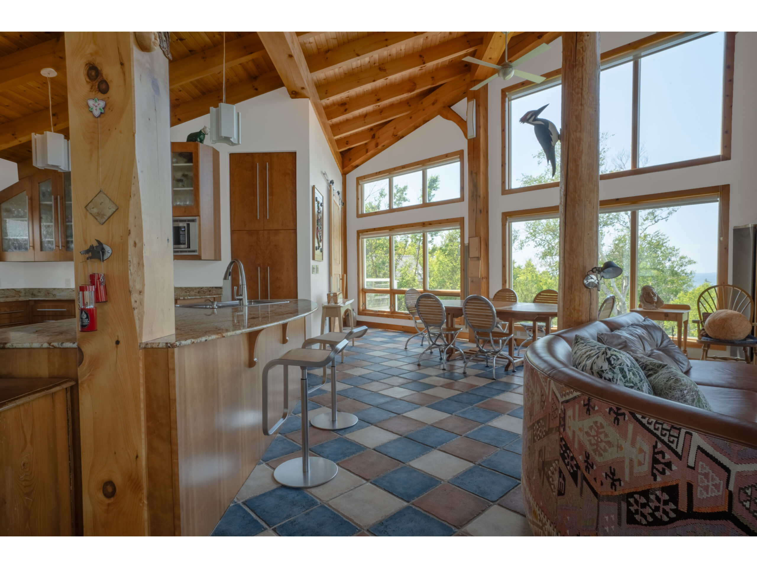 A large, bright, open-concept living area with colourful tiled flooring and wood accents. Big windows reveal green trees and water outside.