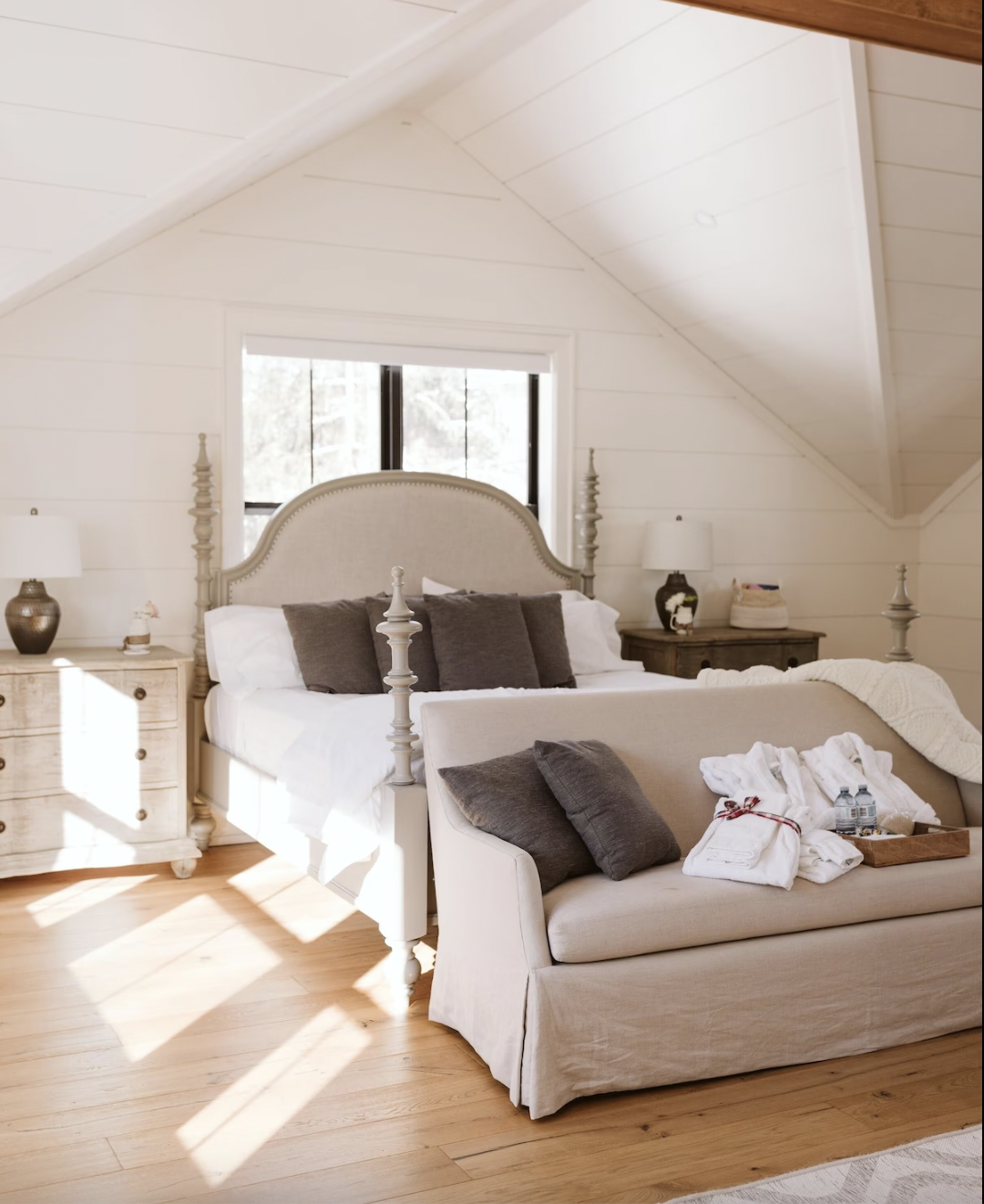 a white room with a large bed with a couch in front of it with pillows and a gift tray on it