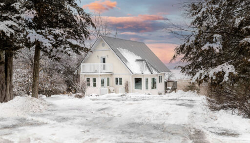 A large home on a snow-covered yard, with large evergreen trees on either side.