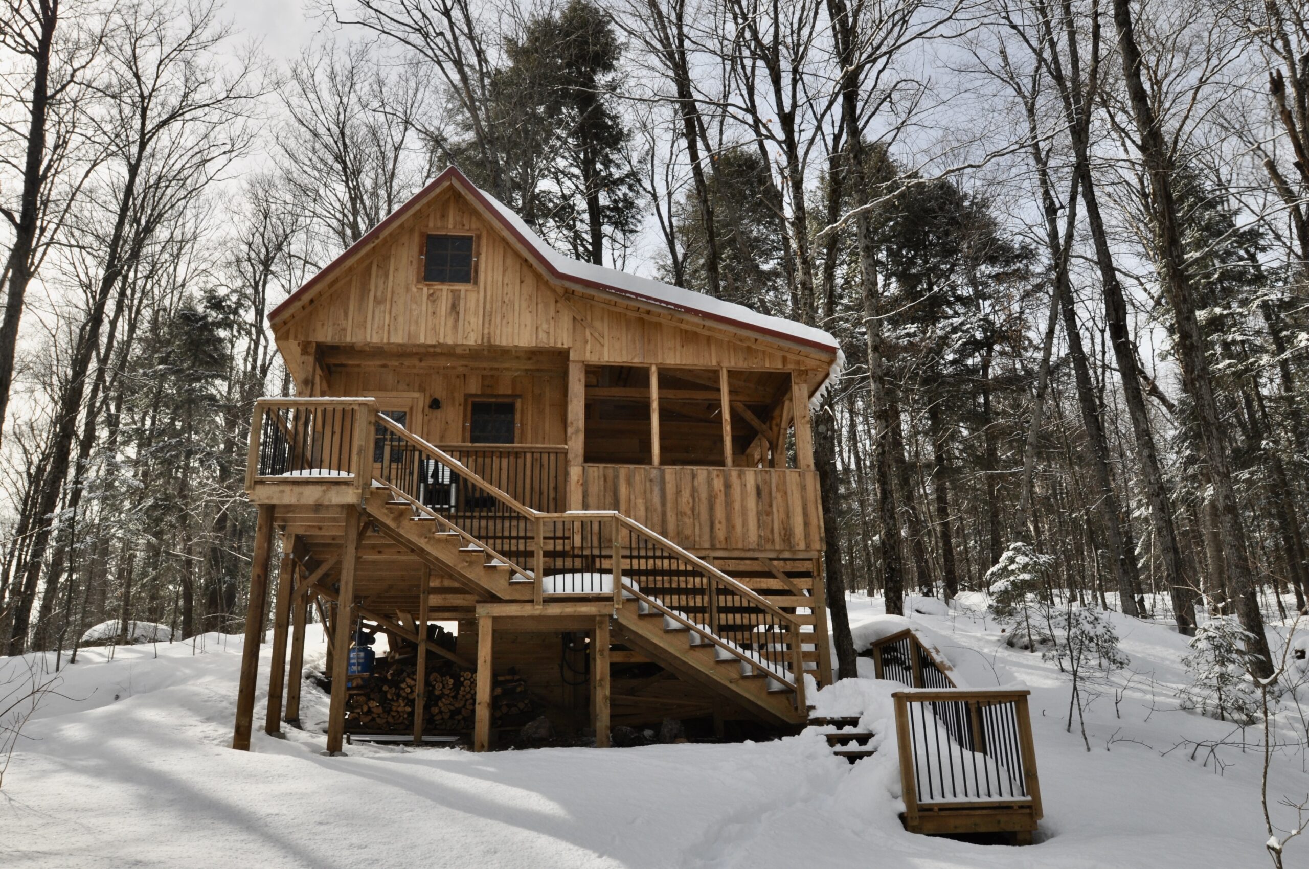 A timber frame cabin with a deck and a screened-in porch sits in a snowy wooded area.