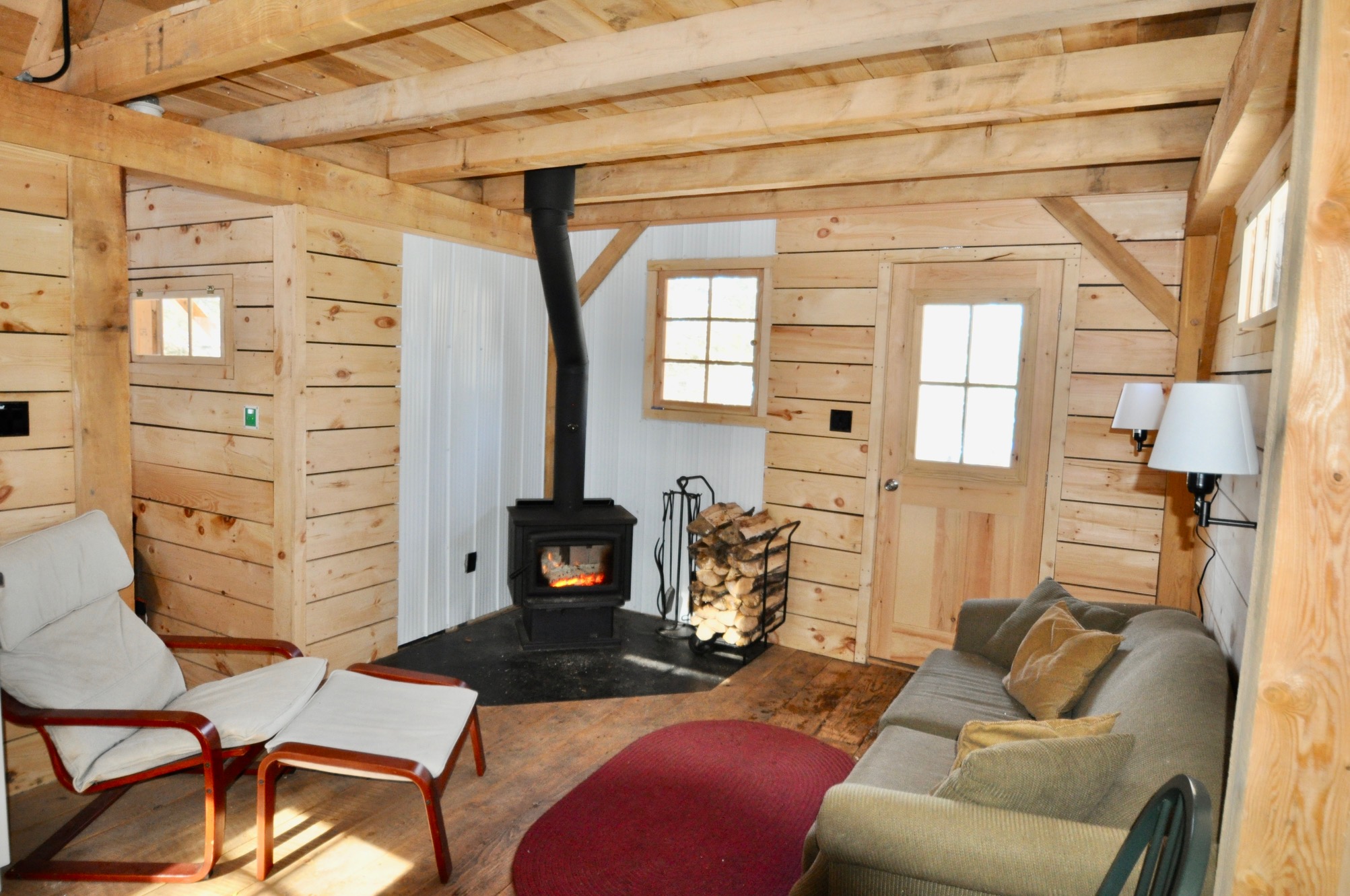 Interior living space of a wood cabin, with a couch and a wood stove. Small windows let in natural light.