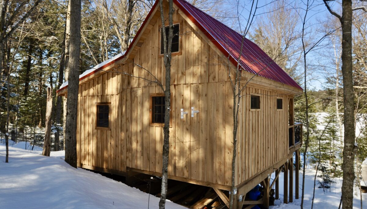 A one and a half story timber frame cabin with a red roof sits in a snowy wooded area.